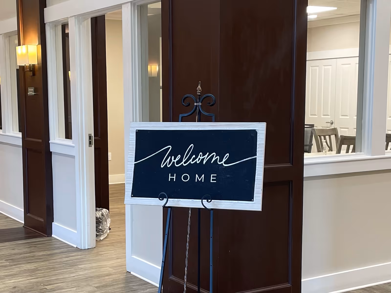 A black and white framed sign on a decorative black metal stand reads 'welcome HOME' in a hallway with wood flooring, white walls, and dark wood paneling. There are windows and chairs visible in the background.