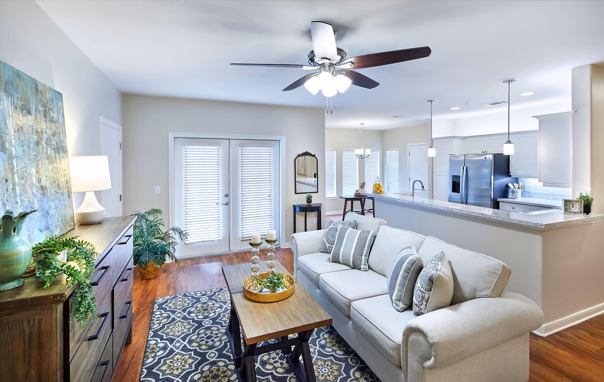 Bright and modern living room with a beige sofa adorned with patterned pillows, a wooden coffee table with decorative items, a patterned rug, a wooden dresser with a lamp and plants, and a ceiling fan with lights. The room opens into a kitchen area with white cabinets, stainless steel refrigerator, and pendant lights over a marble countertop. French doors with blinds lead outside.
