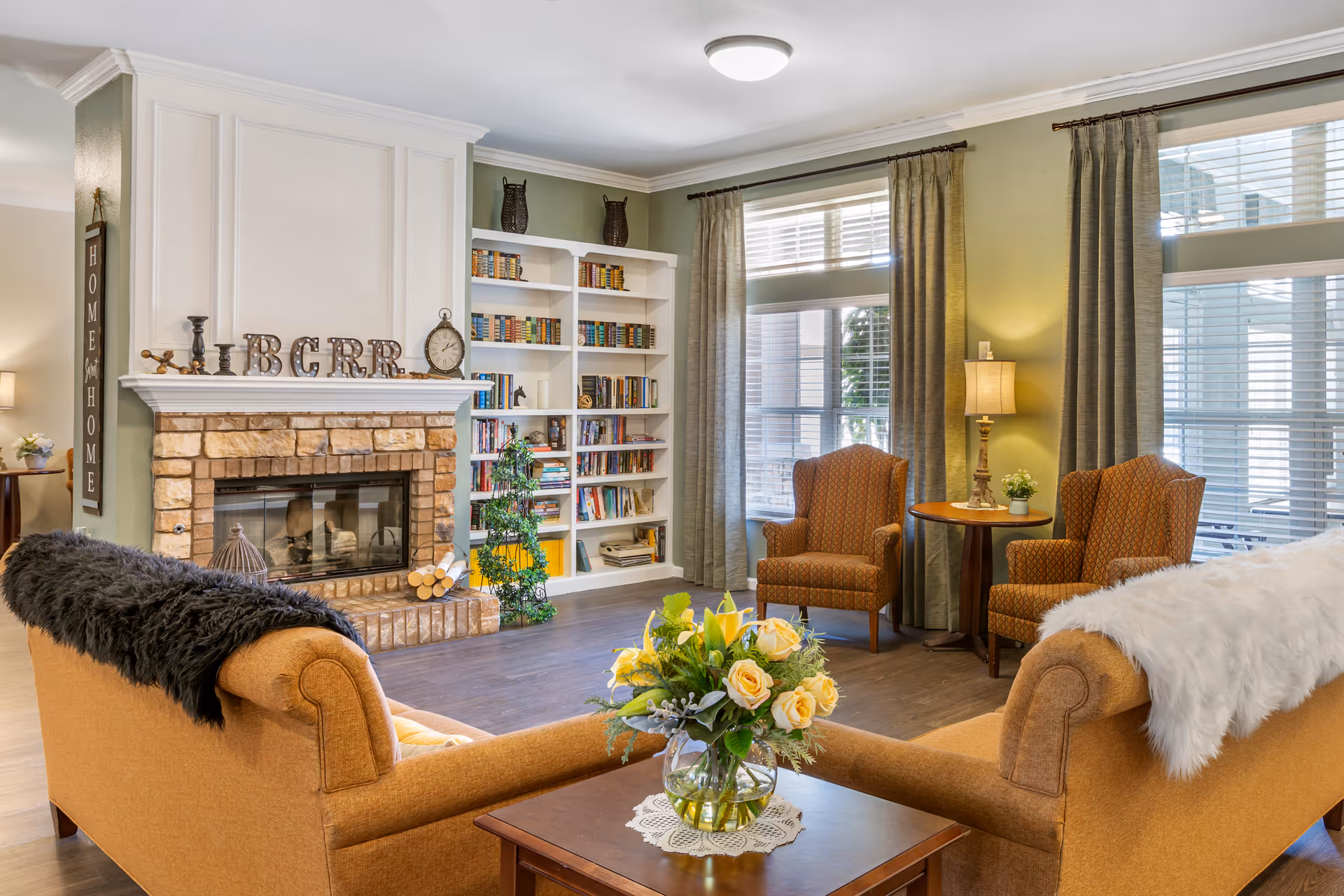 A cozy living room with two orange upholstered armchairs and two matching sofas, one draped with a black throw and the other with a white fur throw. A wooden coffee table with a vase of yellow and white flowers sits in the center. The room features a brick fireplace with decorative items on the mantel, including letters spelling BCRR and a clock. Built-in white bookshelves filled with books and decorative items are on the wall next to large windows with gray curtains, allowing natural light to fill the space.