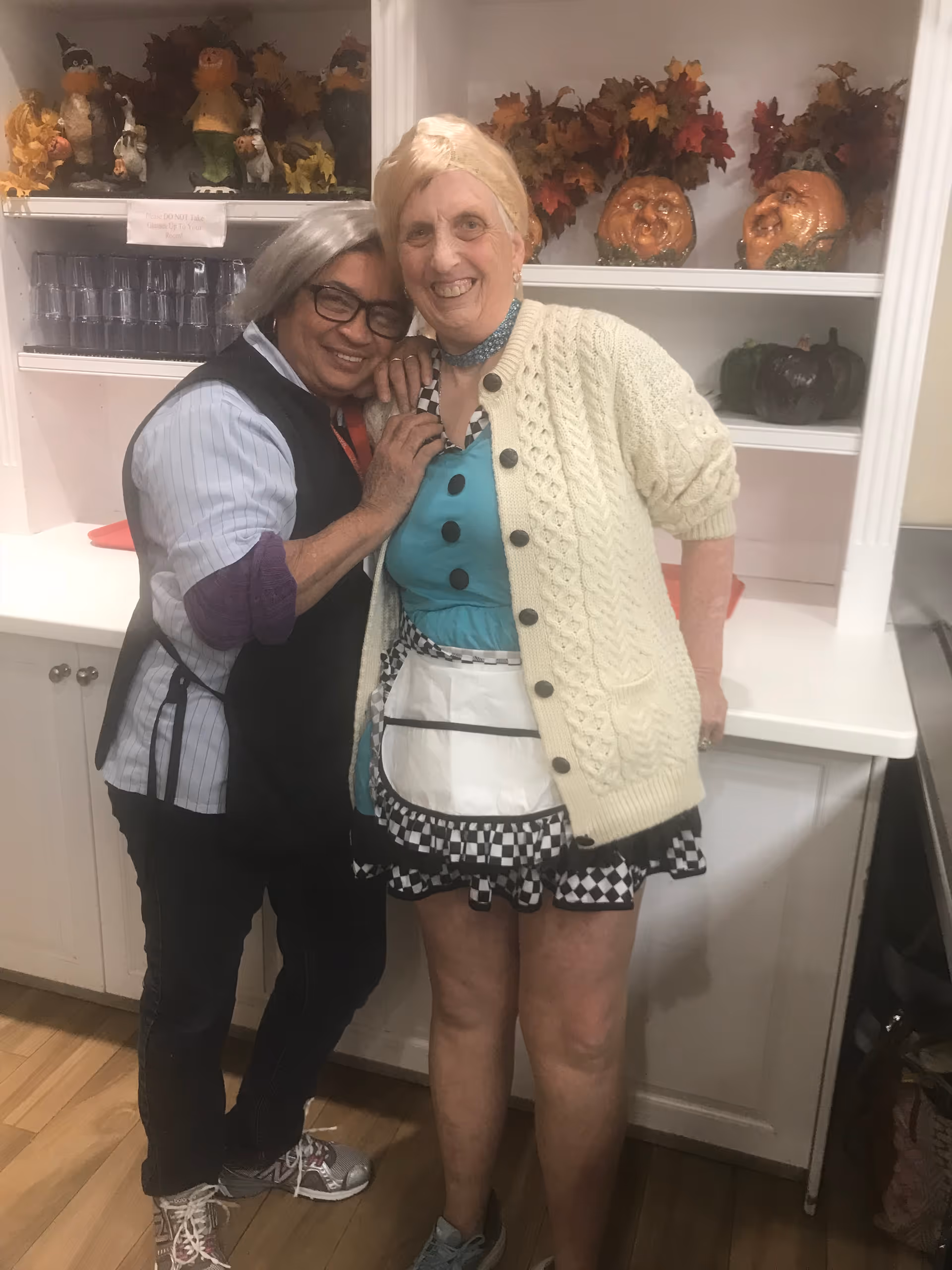 Two smiling residents embrace in a decorated common area with a counter and shelves of fall decorations behind them.