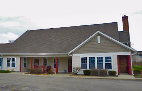 Exterior view of a single-story building with a sloped roof, multiple windows, a chimney, and a covered entrance supported by red pillars. The building is surrounded by a paved area and some small bushes near the walls.