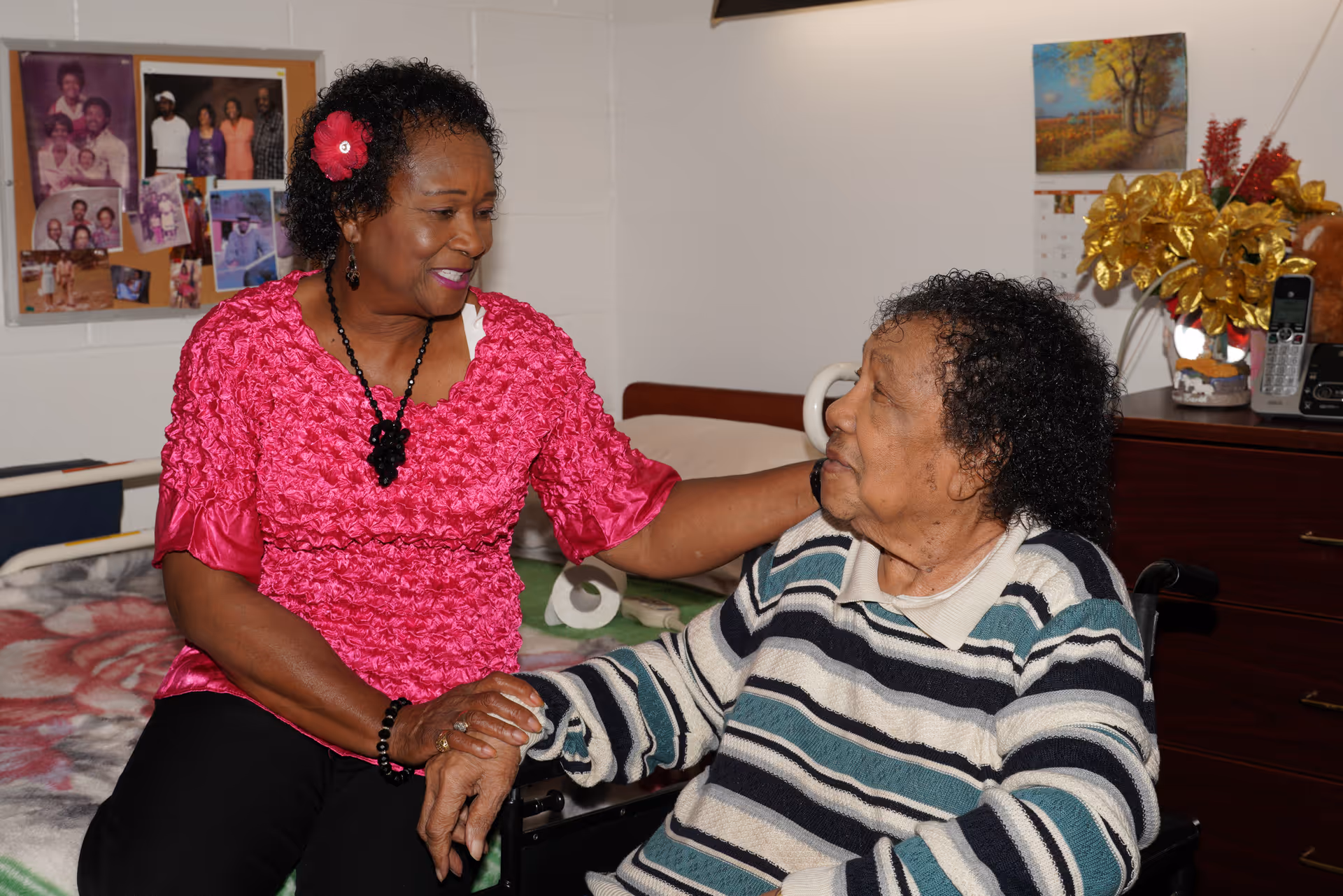 Two elderly women in a cozy bedroom setting. One woman, wearing a bright pink blouse with a flower in her hair, is sitting on a bed and holding hands with the other woman, who is seated in a wheelchair and wearing a striped sweater. The room has a bulletin board with family photos on the wall and a dresser with a telephone and decorative flowers.