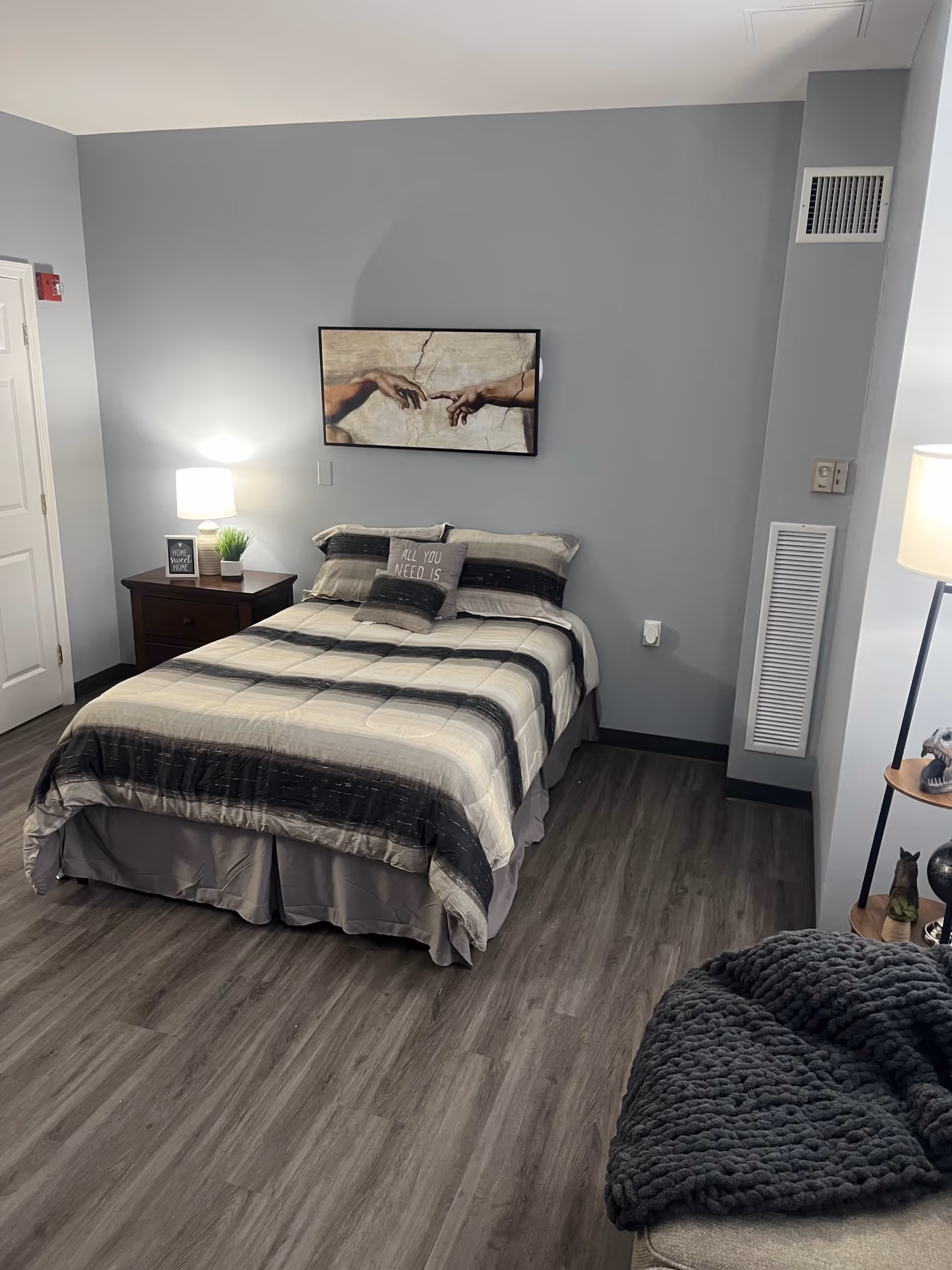 A neatly made bed with a striped comforter and pillows, including one with the text 'ALL YOU NEED IS'. A nightstand with a lamp, a small plant, and a framed sign that says 'HOME SWEET HOME' is next to the bed. Above the bed is a framed artwork depicting two hands reaching towards each other. The room has gray walls and wood flooring, with a cozy blanket draped over a piece of furniture in the foreground.