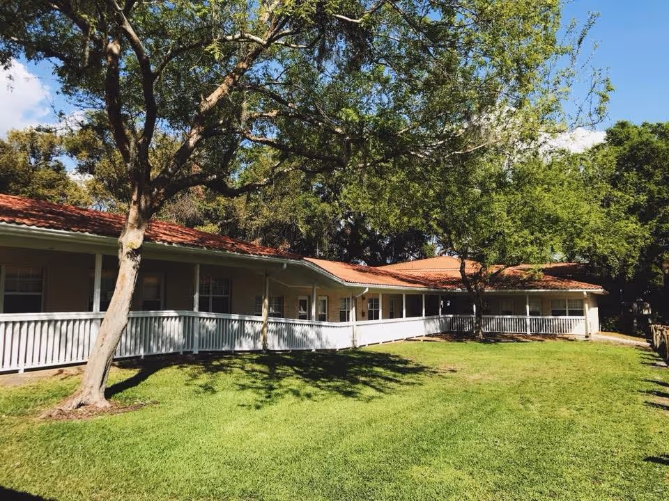 Single-story building with a red-tiled roof and white railing, surrounded by green grass and large trees under a blue sky.