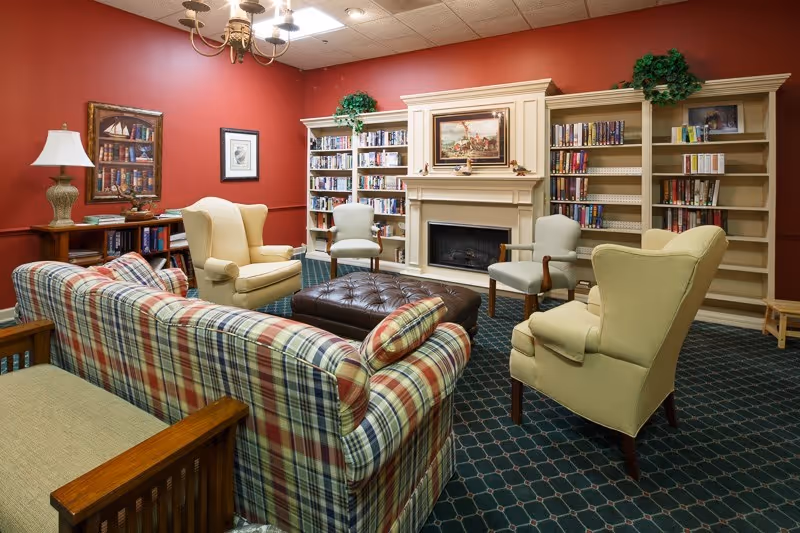 A cozy living room with red walls, a plaid sofa, two beige armchairs, two light green chairs, a large brown ottoman in the center, bookshelves filled with books, a white fireplace with a painting above it, and a chandelier hanging from the ceiling.