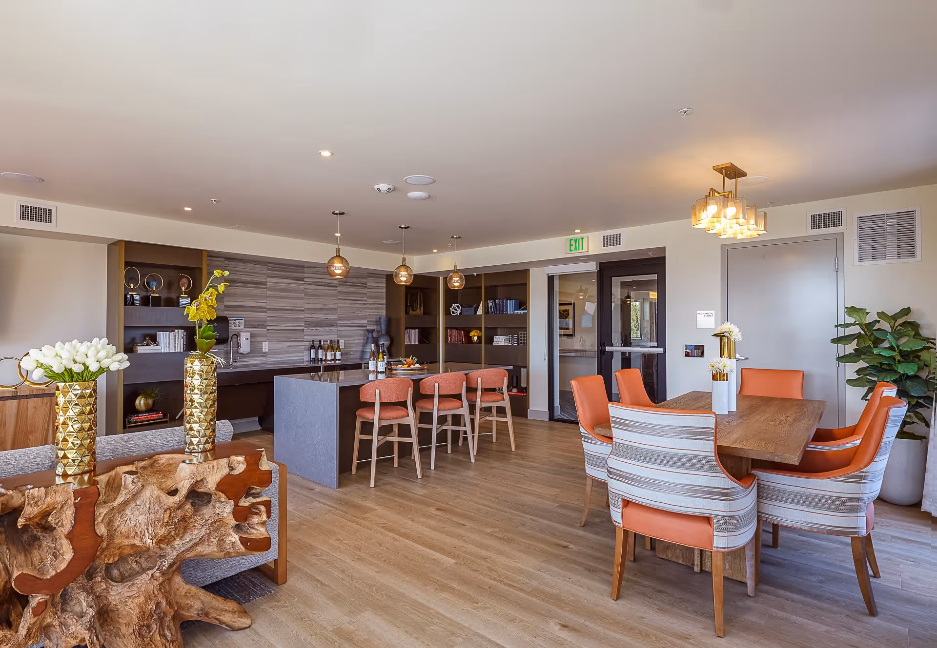 A modern and spacious dining and kitchen area in a senior living facility. The room features a wooden dining table with six orange and striped upholstered chairs, a kitchen island with four orange bar stools, pendant lights hanging above the island, and decorative vases with flowers on the tables. The background includes built-in shelves with books and decorative items, a door, and a large plant in the corner.