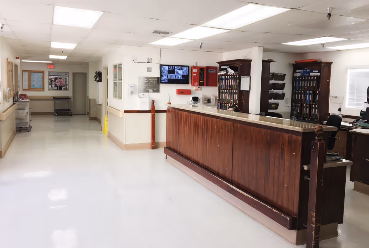 A clean and well-lit reception area inside a senior care facility with a long wooden front desk, office chairs, and organized shelves behind the desk. The hallway extends to the left with white walls, a bulletin board, and a cart. Security monitors and fire safety equipment are mounted on the wall behind the desk.