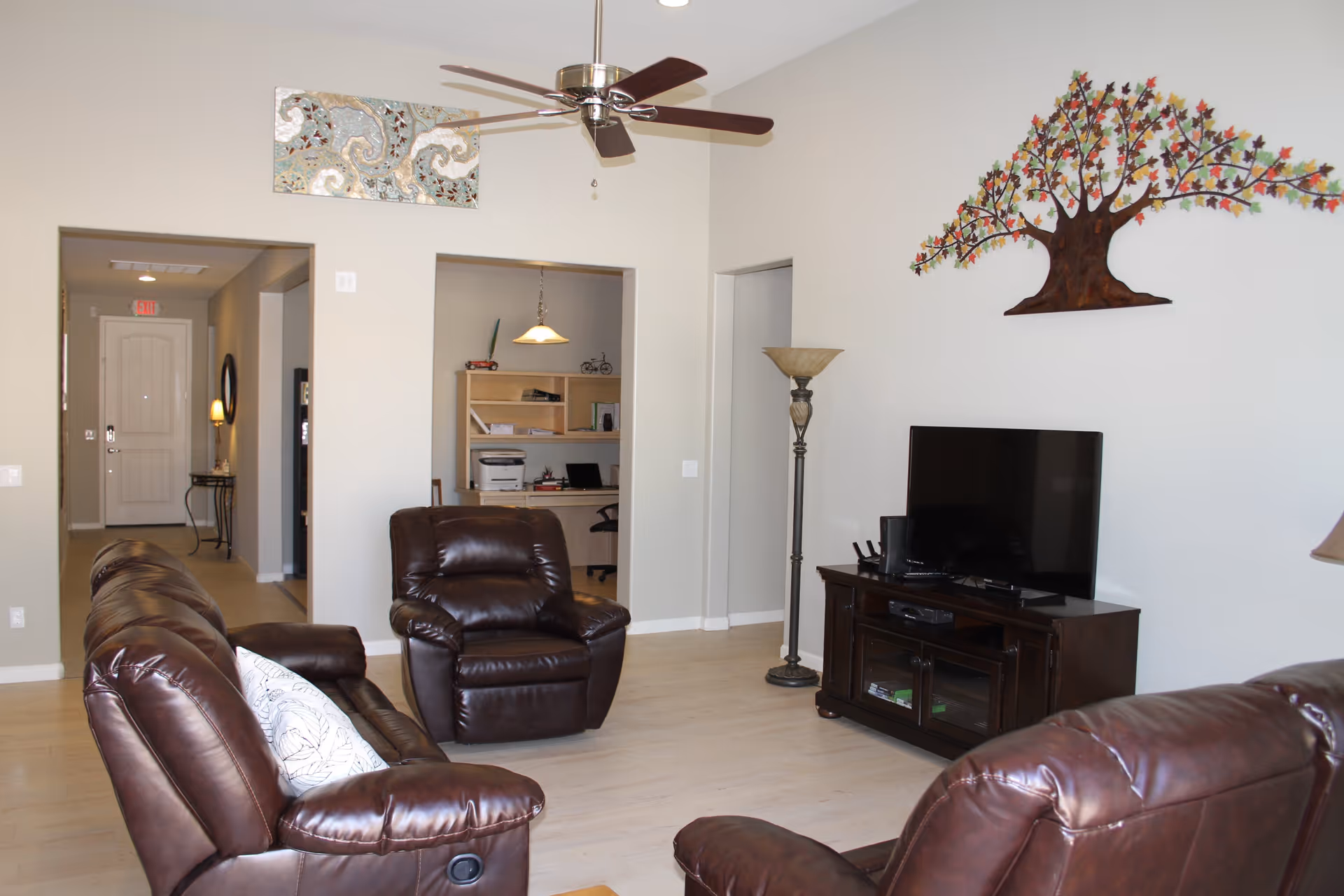 A living room with brown leather sofas and a recliner arranged around a TV on a wooden stand. The room has light-colored wooden flooring and white walls. A ceiling fan with wooden blades is mounted on the ceiling. There is a decorative metal tree wall art above the TV and a floor lamp next to the TV stand. In the background, there is an open doorway leading to a small office area with a desk, chair, and shelves.