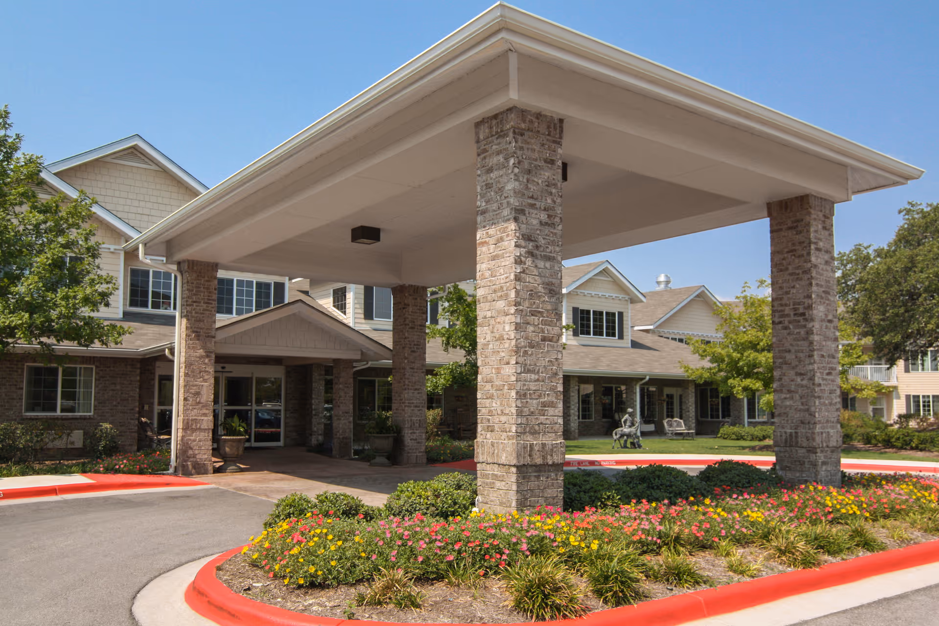 Entrance of Asher Point Independent Living of Round Rock showing a covered drop-off area supported by brick pillars, surrounded by landscaped flower beds with yellow and pink flowers, and a multi-story building with beige siding and multiple windows in the background under a clear blue sky.