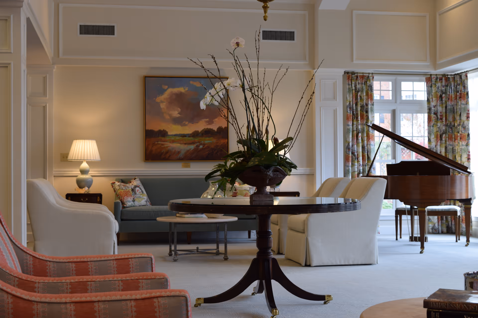 Elegant living room with cream walls, floral curtains, grand piano, round table, and comfy seating.