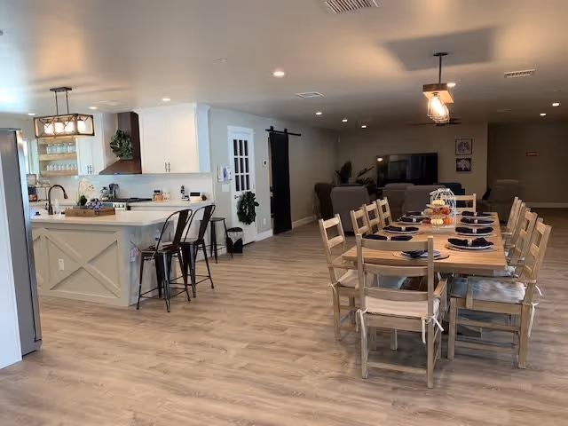 A spacious, modern open-concept interior featuring a kitchen area with white cabinets, a kitchen island with bar stools, and a dining area with a wooden table set for eight with navy blue napkins. The room has light wood flooring, neutral-colored walls, and ceiling lights. In the background, there is a living area with armchairs and a TV.