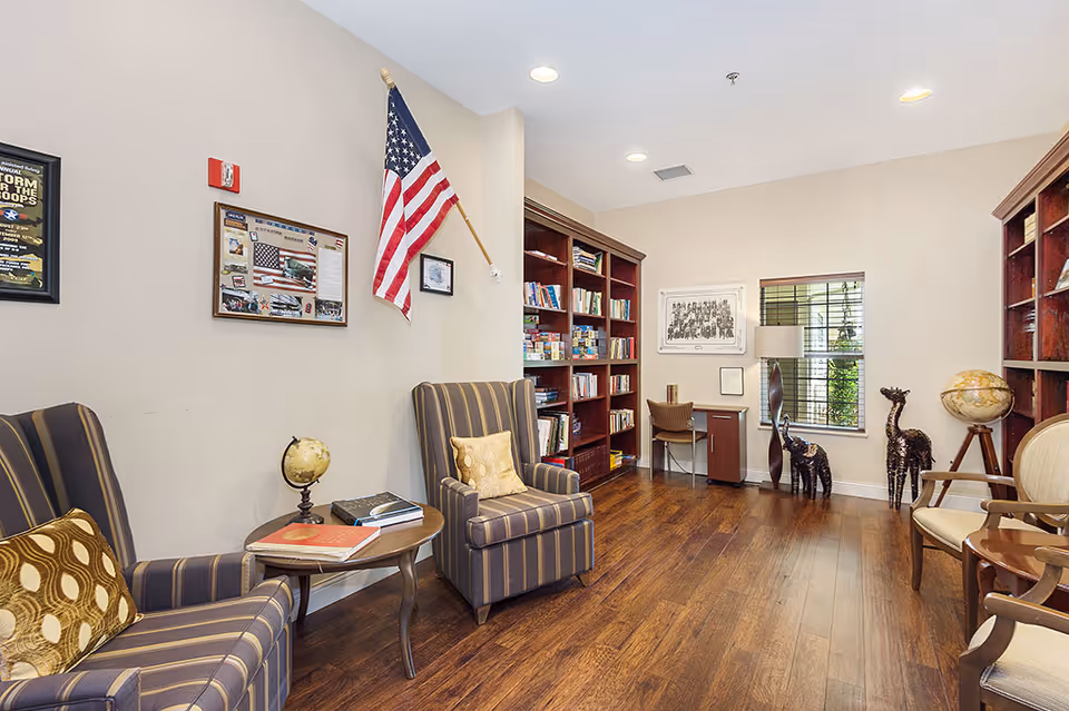 Cozy reading room with striped armchairs, bookshelves, a small desk, decorative globes and giraffe sculptures, and an American flag on the wall.