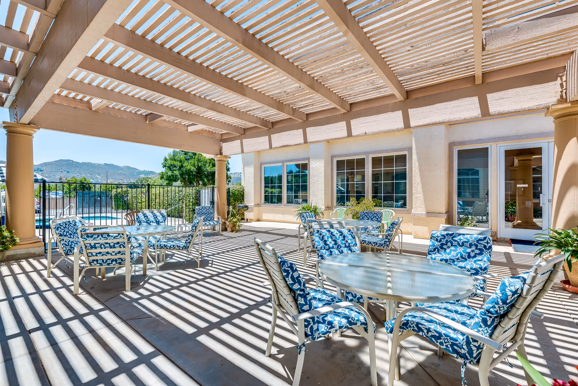 Shaded outdoor patio with round glass tables and blue patterned cushioned chairs under a pergola, adjacent to a pool and building.