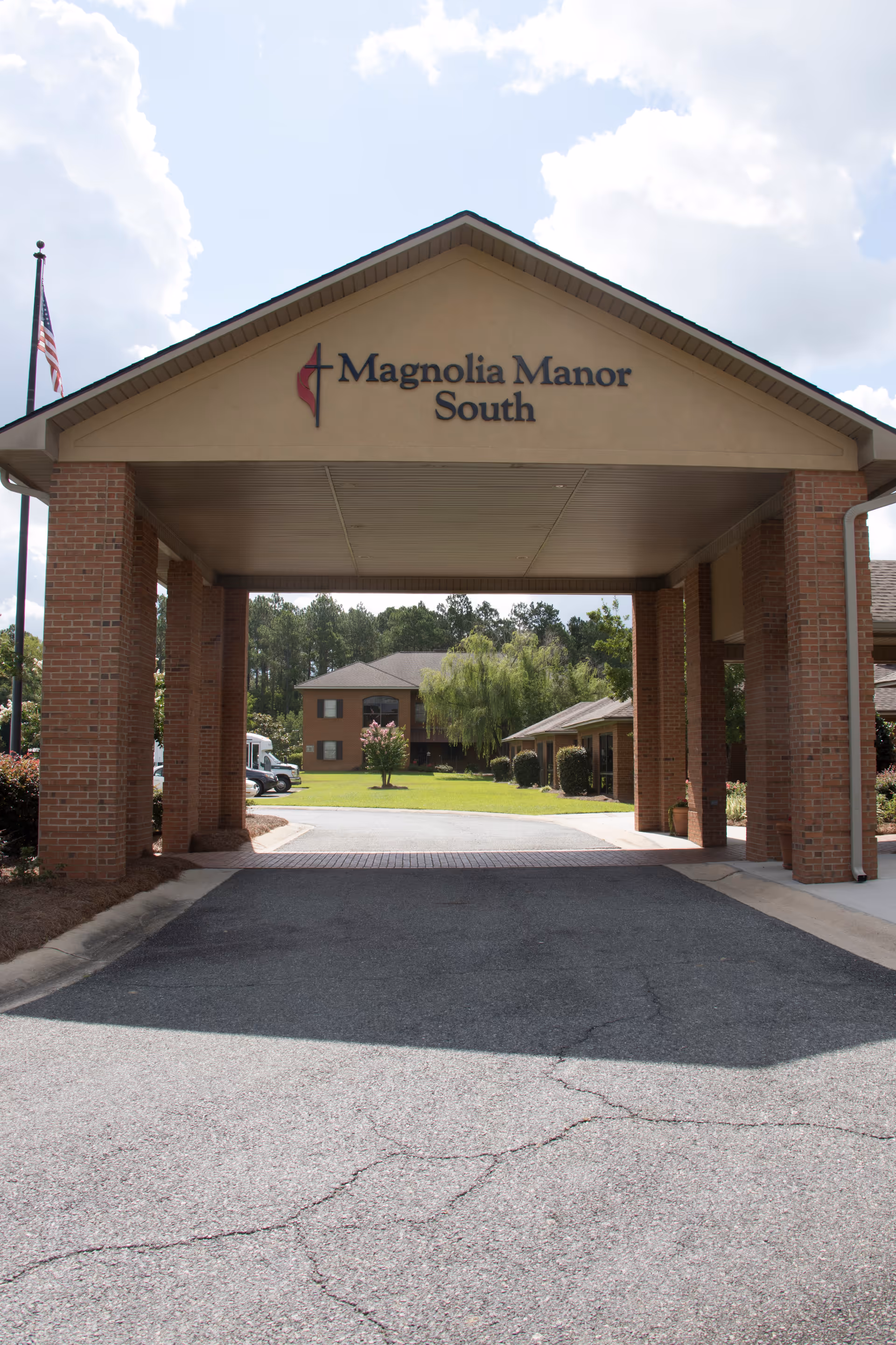 Entrance canopy of Magnolia Manor South, a senior living facility, with brick pillars and a driveway leading to the building surrounded by greenery and trees under a partly cloudy sky.
