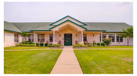 Front exterior of a single-story brick building with a central covered entrance, walkway, and manicured lawn.
