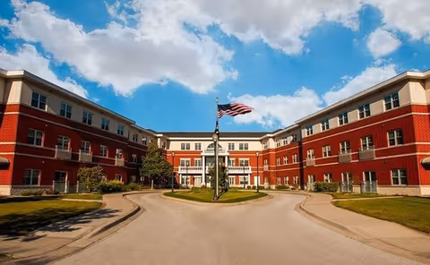 Exterior view of a three-story senior living facility building with red brick and beige accents under a partly cloudy blue sky. A circular driveway with a flagpole flying the American flag is in front of the building.