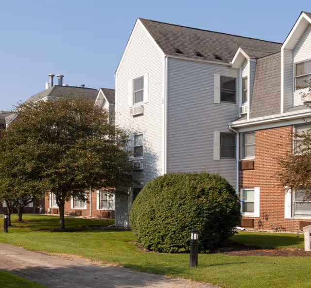 Exterior view of a senior living facility building with white siding and brick walls, surrounded by green grass, trees, and bushes under a clear blue sky.