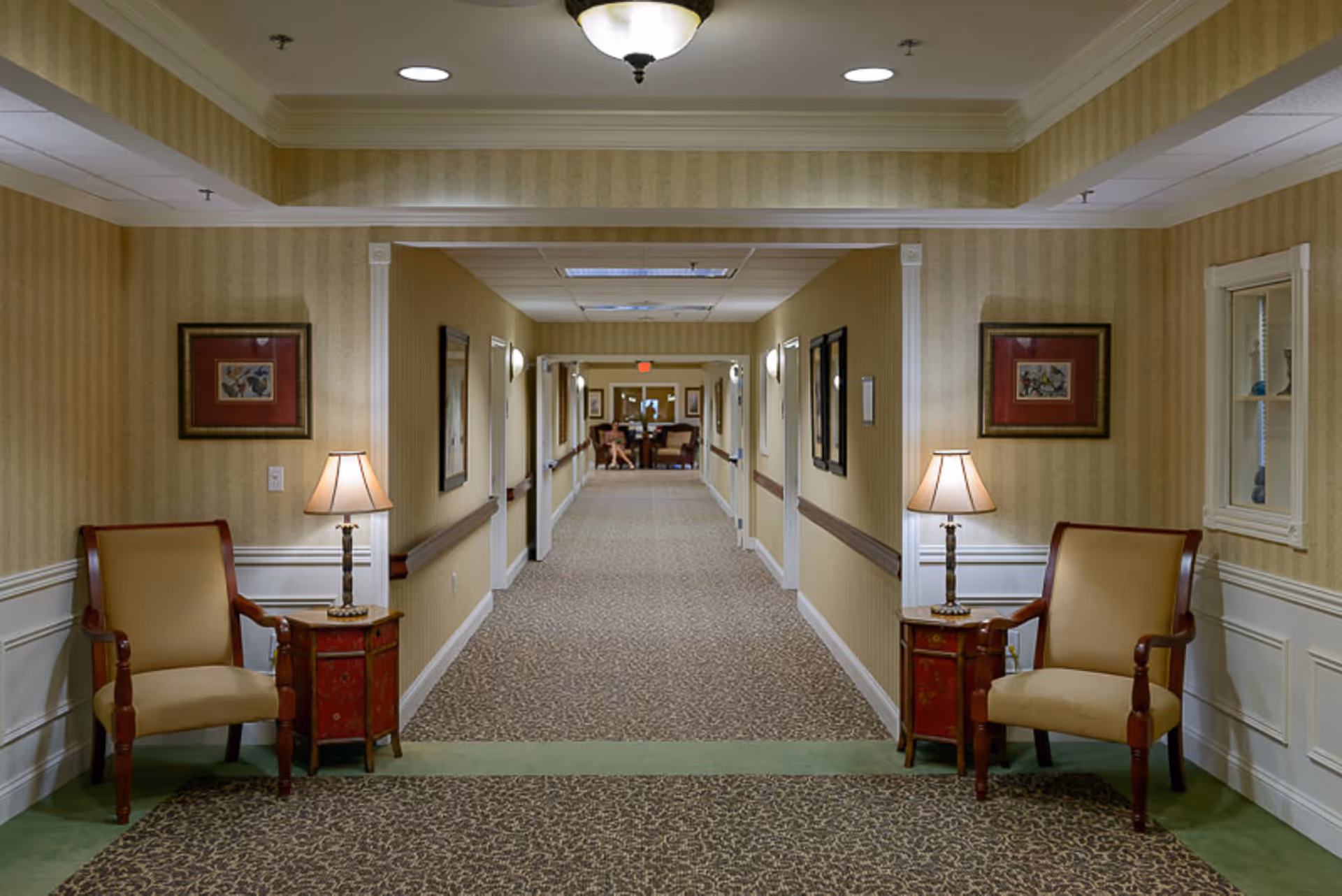 A long, carpeted hallway in a senior living facility with beige striped wallpaper and white wainscoting. Two beige armchairs with wooden frames are placed on either side of the hallway entrance, each accompanied by a small red side table with a lamp. The hallway is well-lit with ceiling lights and wall sconces, and framed artwork is hung along the walls. At the far end of the hallway, there is a seating area with two chairs and a table.