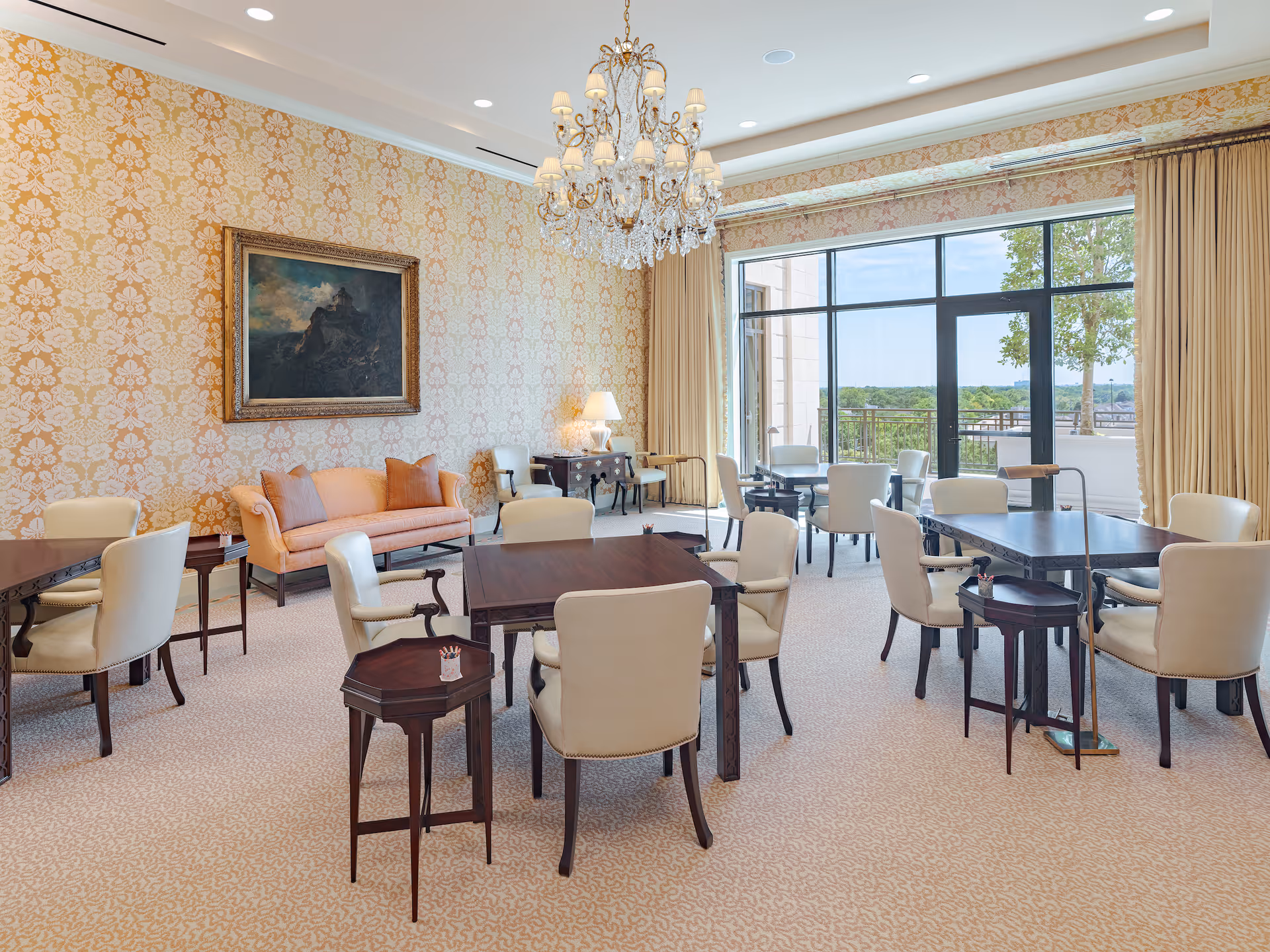 A bright and elegant dining room with multiple dark wood tables and cream upholstered chairs. The room features a large window with a view of trees and a balcony outside. There is a peach-colored sofa against a wall with ornate wallpaper, a framed painting above it, and a crystal chandelier hanging from the ceiling.