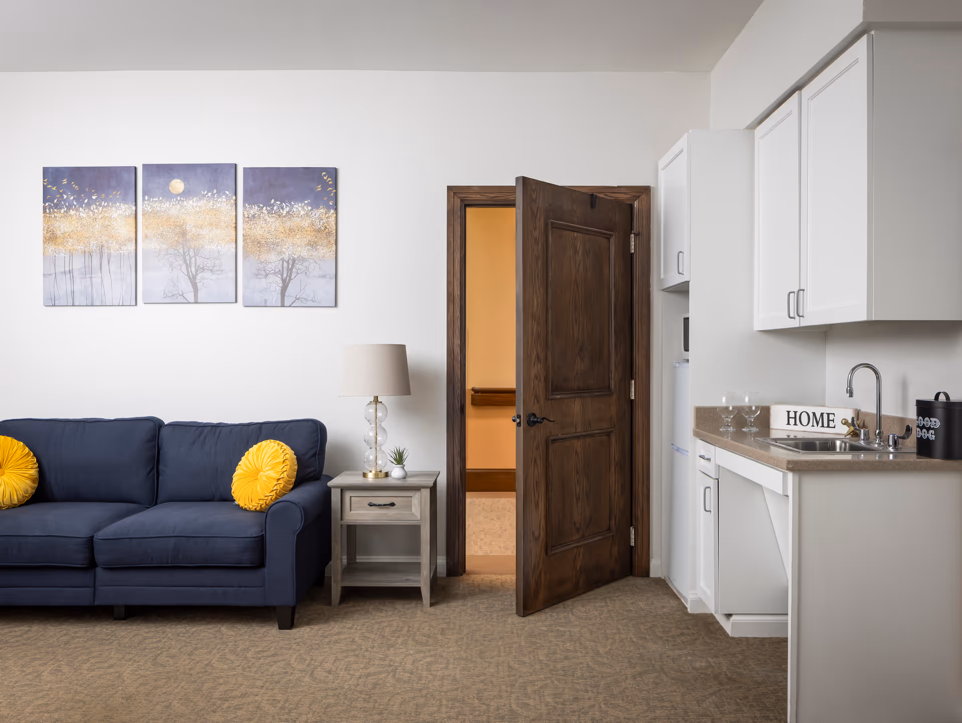 A cozy living area featuring a navy blue couch with two round yellow pillows, a small wooden side table with a lamp and a small plant, a wooden door slightly ajar revealing a hallway, and a kitchenette with white cabinets, a sink, and a countertop with two wine glasses and a decorative HOME sign.