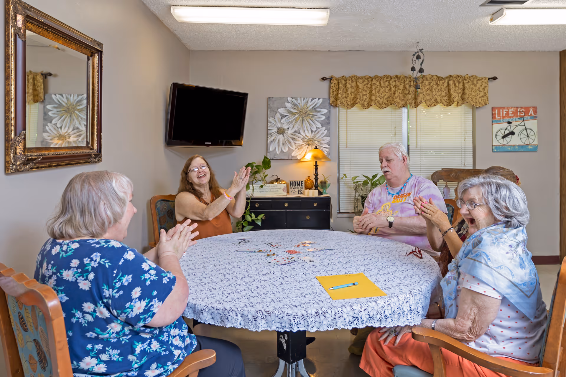 A group of five elderly people sitting around a round table covered with a white lace tablecloth in a cozy room. They appear to be engaged in a card game and are clapping and smiling. The room has a wall-mounted TV, a large mirror, decorative wall art, a window with blinds and a valance, and a small black cabinet with a lamp and plants on it.