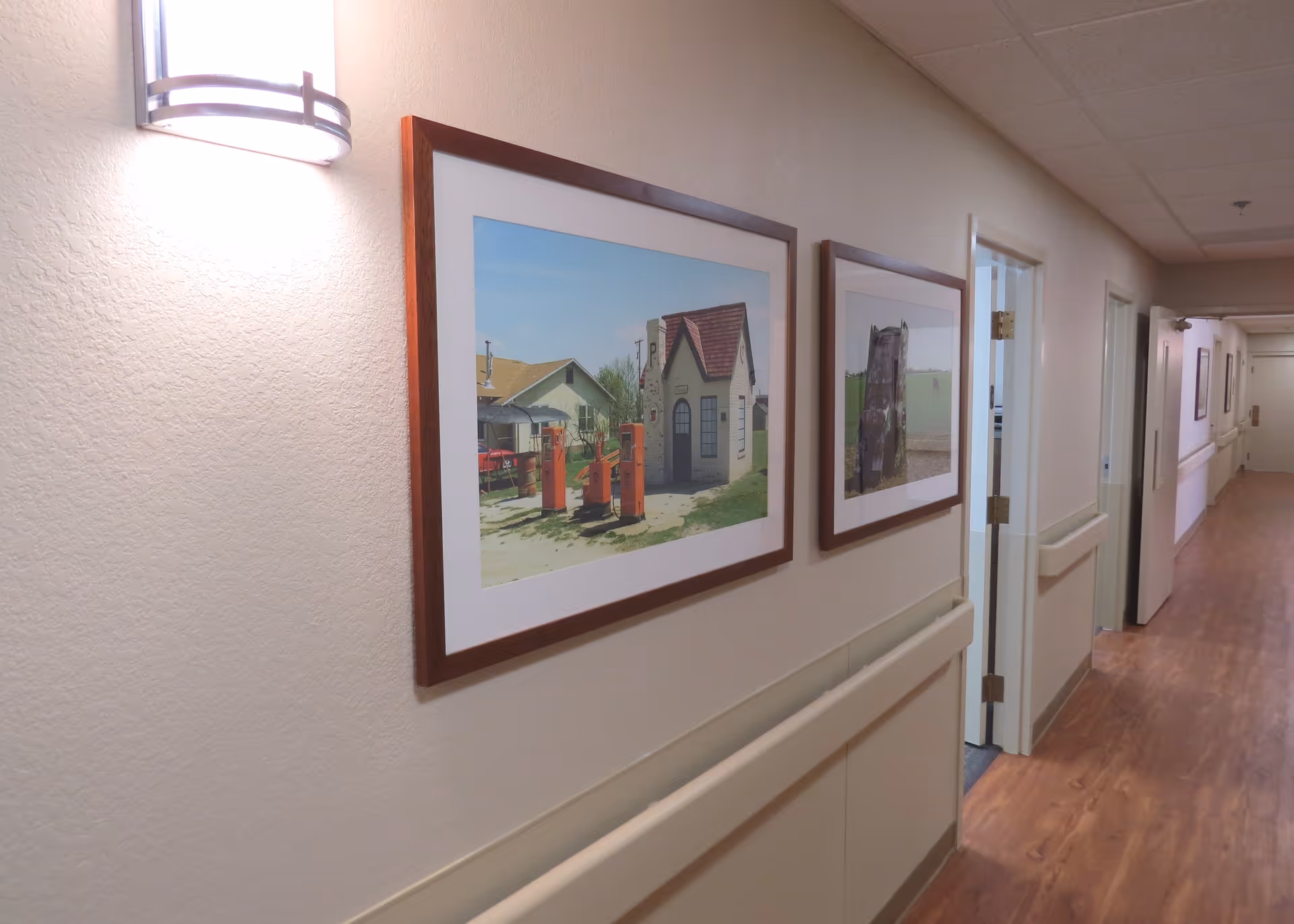 A well-lit interior hallway with framed artwork, handrails, doors, and wood-look flooring in a nursing facility.