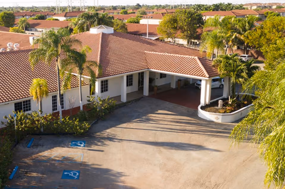 Exterior view of a single-story building with a red tiled roof and white walls, surrounded by palm trees and greenery. There is a covered entrance with columns and a small landscaped area with plants. The parking area in front includes two handicap parking spaces.