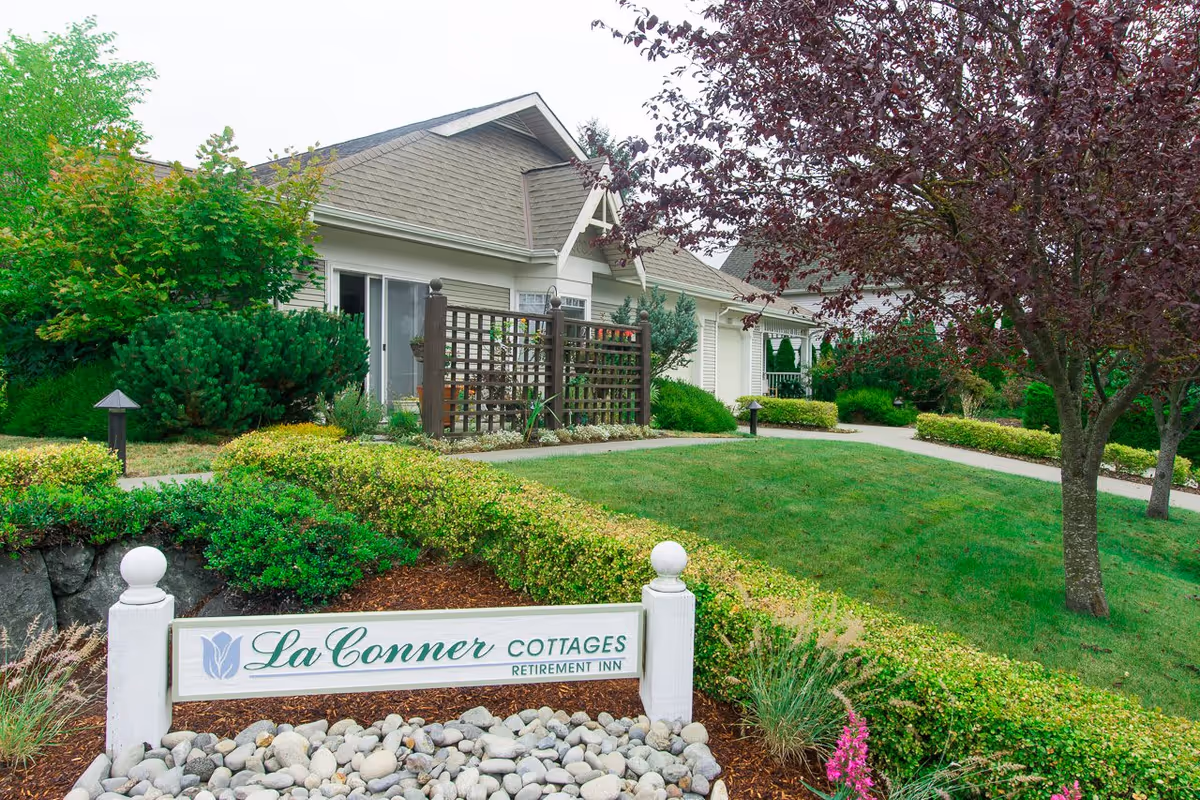 Exterior view of La Conner Cottages Retirement Inn showing a well-maintained garden with green bushes, trees, and a lawn. The building has a light-colored exterior with a sloped roof and a wooden lattice fence near the entrance.