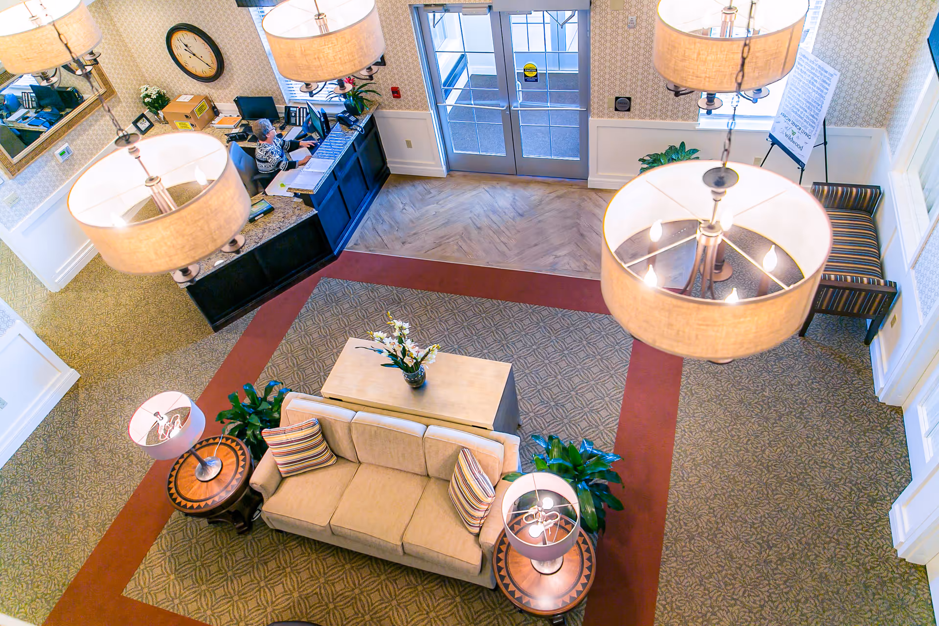 Overhead view of a senior living facility lobby with a beige sofa, two side tables with lamps, a coffee table with a flower vase, potted plants, a reception desk with a staff member working, large hanging light fixtures, and double glass doors leading outside.