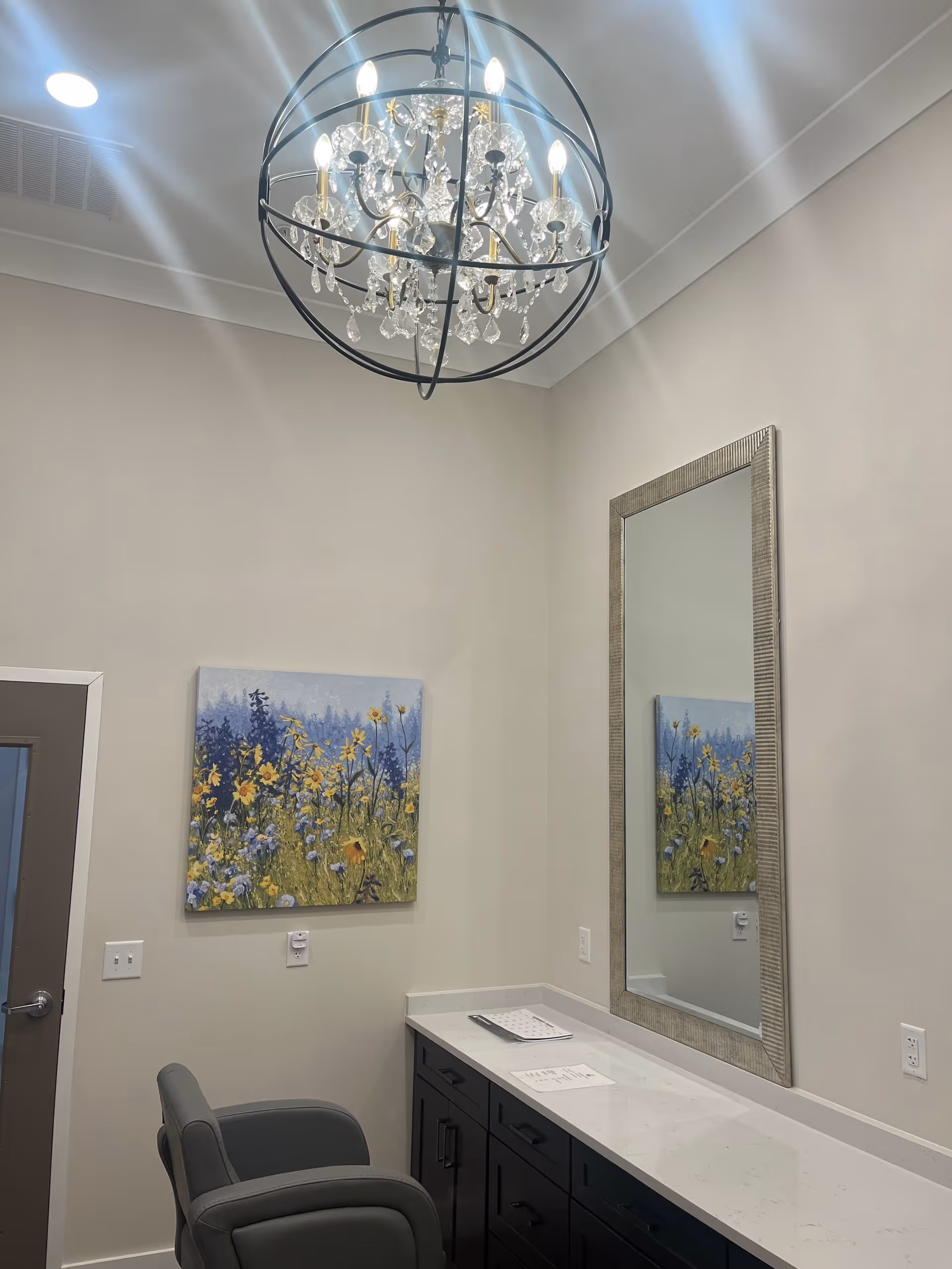 A well-lit interior room with a modern chandelier hanging from the ceiling. Below is a long countertop with dark cabinets and a large rectangular mirror mounted on the wall. A gray cushioned chair is positioned in front of the counter. On the wall, there is a colorful painting of wildflowers and trees. The room has light-colored walls and a door with a silver handle on the left side.