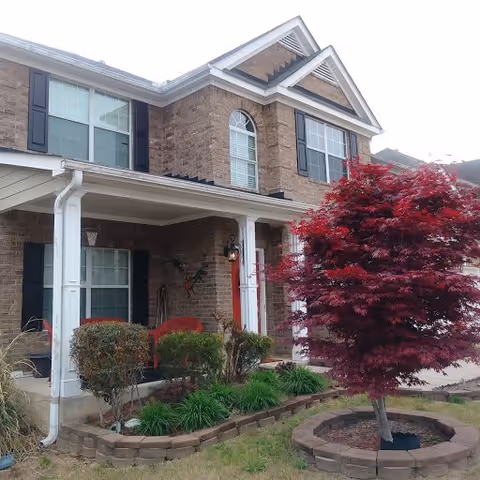 Front of a two-story brick building with a covered porch, shrubs and a red-leaved ornamental tree in the yard.