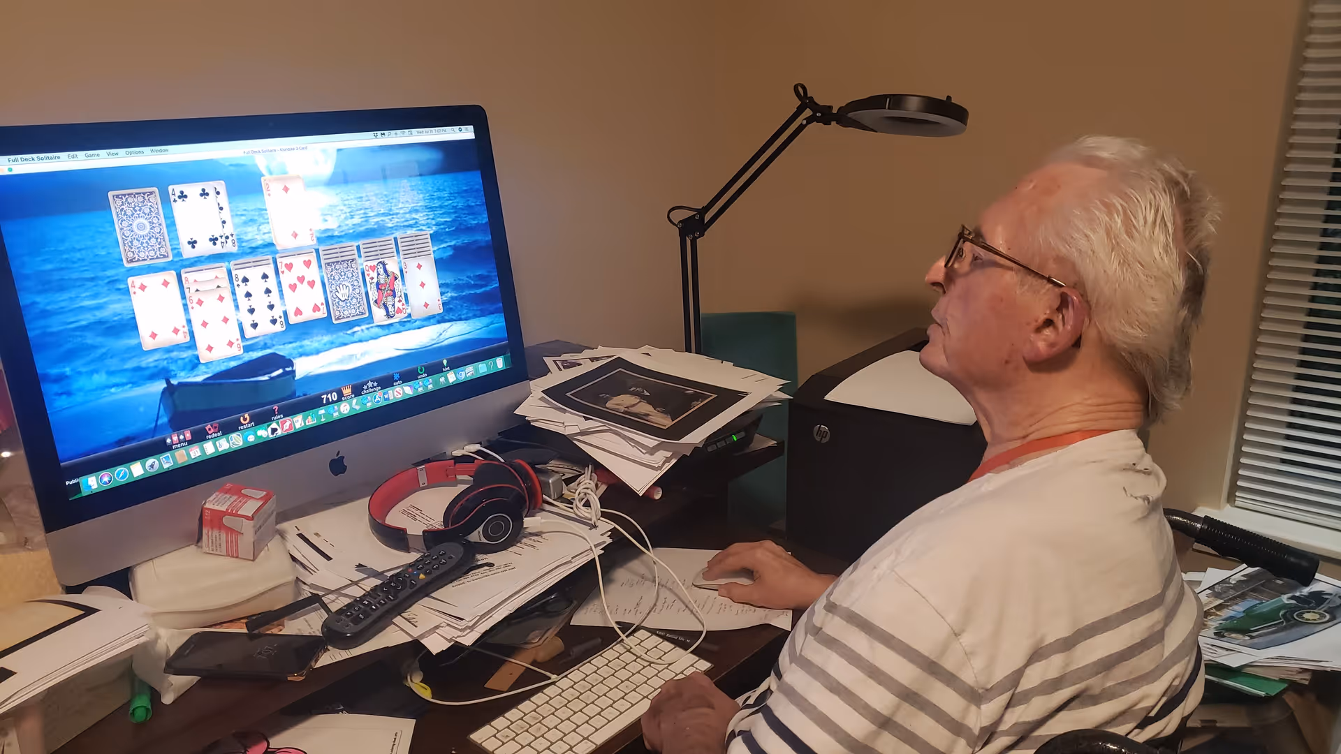 An elderly man with white hair and glasses is sitting at a cluttered desk playing a solitaire card game on a large iMac computer. The desk is filled with papers, a remote control, headphones, and other miscellaneous items. A desk lamp and a printer are also visible in the background.