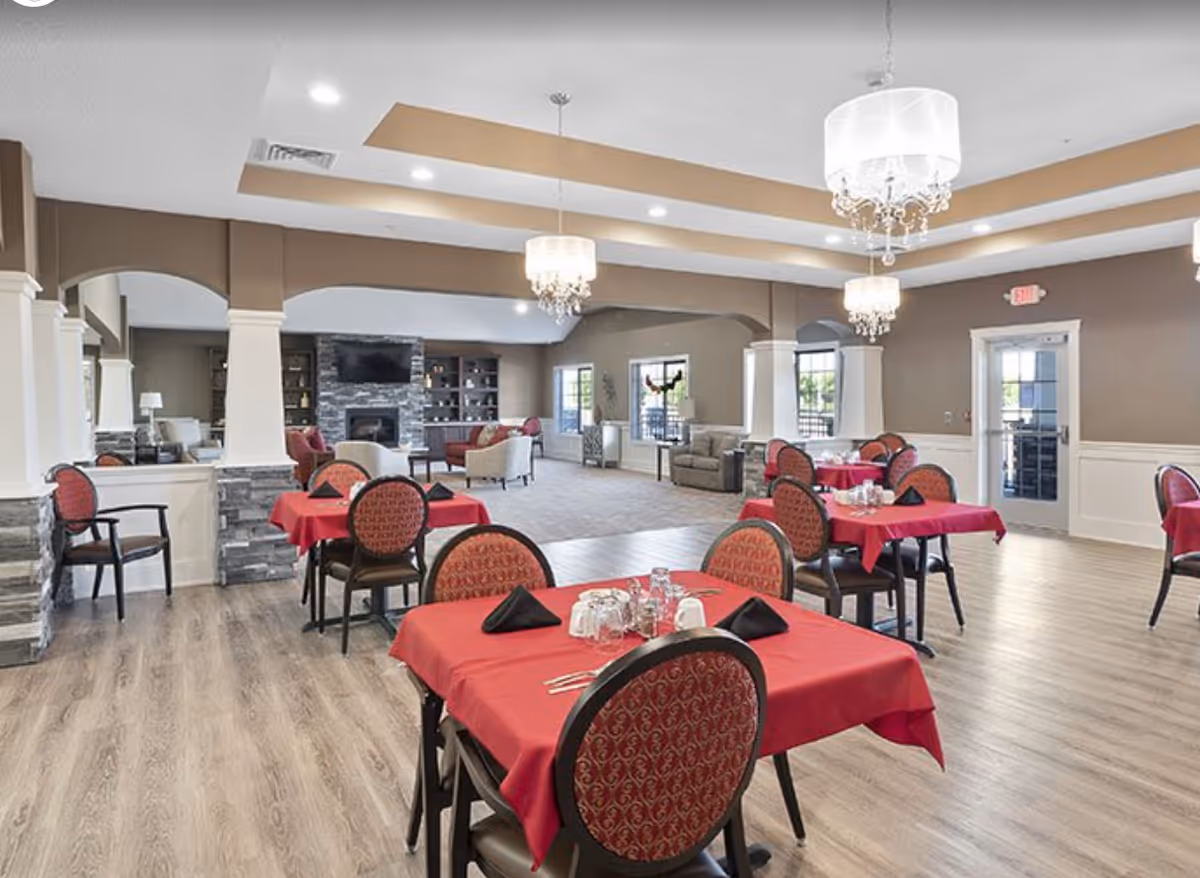 A spacious dining room with several tables covered in red tablecloths and set with black napkins, glasses, and silverware. The room features wood flooring, beige walls with white wainscoting, and elegant chandeliers hanging from the ceiling. In the background, there is a cozy seating area with armchairs and a stone fireplace with a mounted TV above it. Large windows and glass doors allow natural light to enter the room.