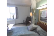 A simple bedroom in a nursing facility with a bed covered in a blue quilt, a pillow resting on the bed, a chair near the window, a curtain partially drawn around the bed, and a framed picture on the wall.