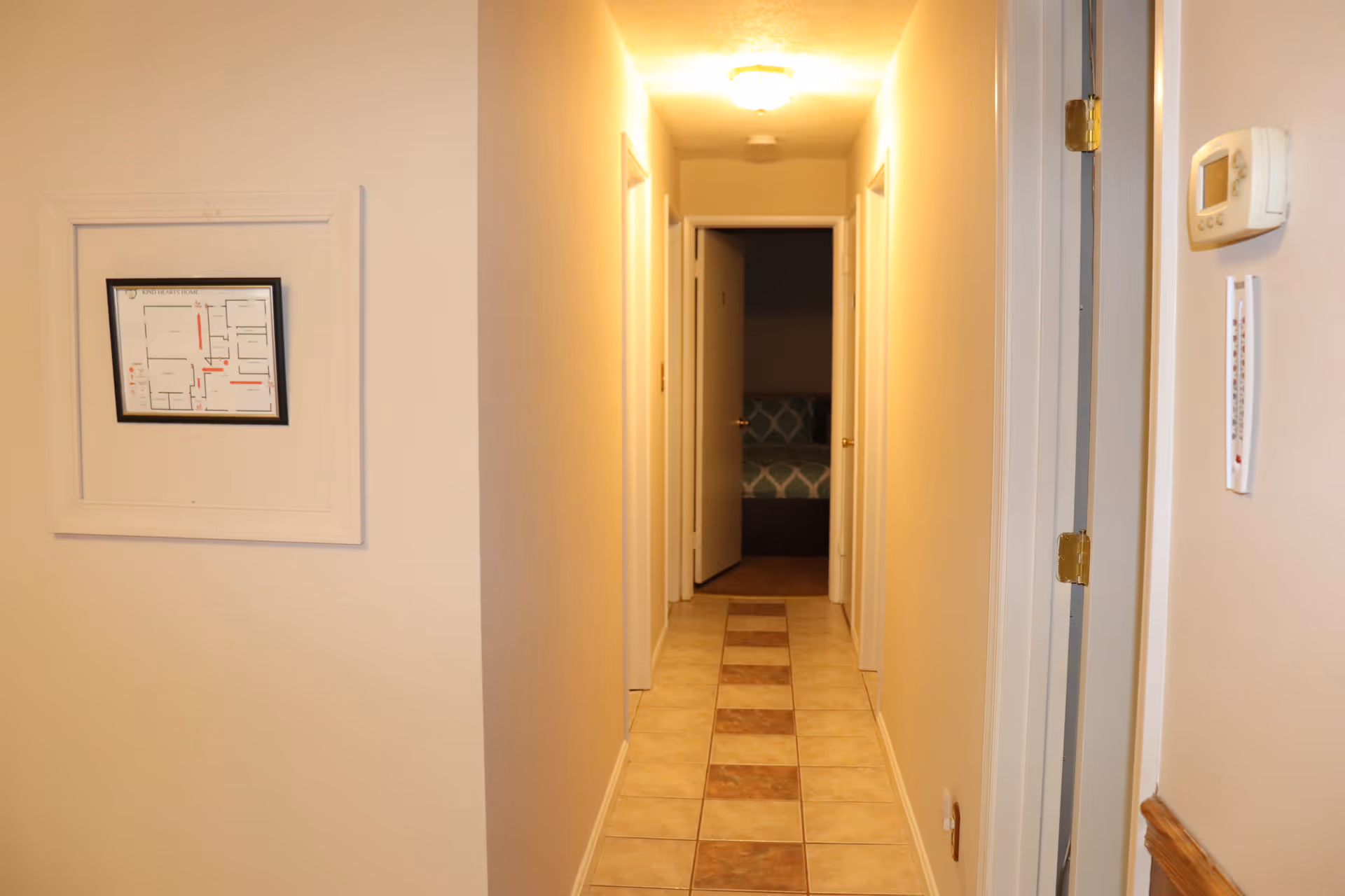 A narrow hallway with beige walls and tiled floor featuring a pattern of alternating light and dark tiles. There are several closed white doors along the hallway, and one door at the end is open, revealing a bedroom with a bed covered in a blue and white patterned bedspread. A framed floor plan is mounted on the left wall, and a thermostat and thermometer are visible on the right wall.