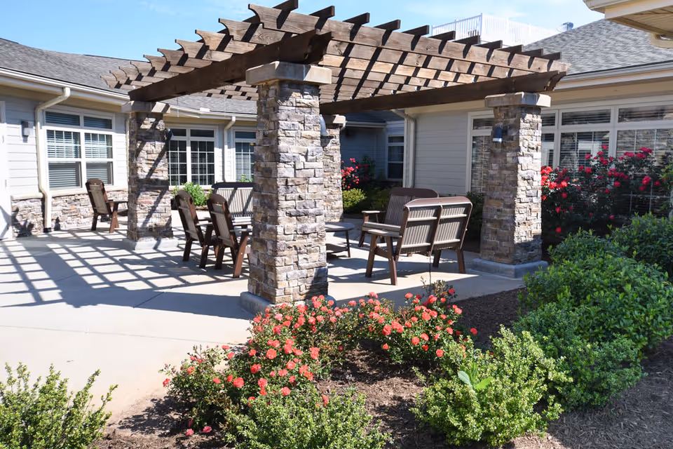 Outdoor patio area at Oakview Park featuring a wooden pergola supported by stone pillars, with several wooden chairs and benches arranged underneath. Surrounding the patio are well-maintained flower beds with blooming red flowers and green shrubs, adjacent to the building with multiple windows.