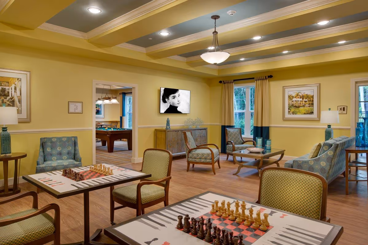 A cozy and well-lit common area in a senior living facility featuring two tables with chess boards and chairs, comfortable armchairs and sofas, a wooden coffee table, framed artwork on yellow walls, and a flat-screen TV displaying a black and white portrait. In the background, there is an adjacent room with a pool table and additional seating.
