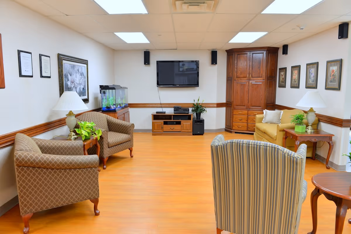 A cozy living room area with wooden flooring, featuring a wall-mounted flat-screen TV, a wooden entertainment center, a fish tank, two patterned armchairs, a striped armchair, a yellow loveseat, side tables with lamps and plants, and framed artwork on the walls.