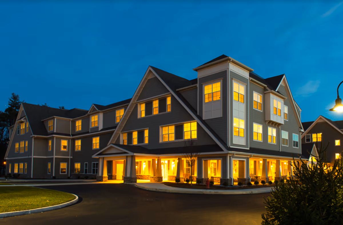 A large multi-story senior living building at dusk with warmly lit windows and an illuminated covered entrance.