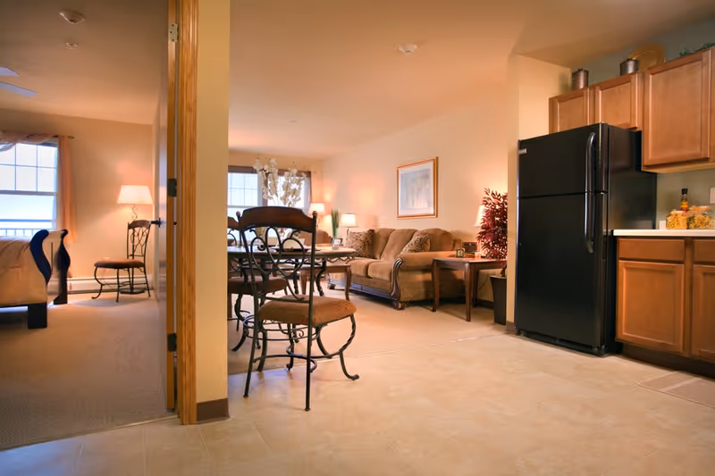 View of a cozy assisted living apartment showing a kitchen area with wooden cabinets and a black refrigerator on the right, a dining table with chairs in the center, and a living room with a sofa, side table, lamp, and framed artwork on the wall. To the left, a bedroom with a bed, chair, and lamp is partially visible through an open doorway.