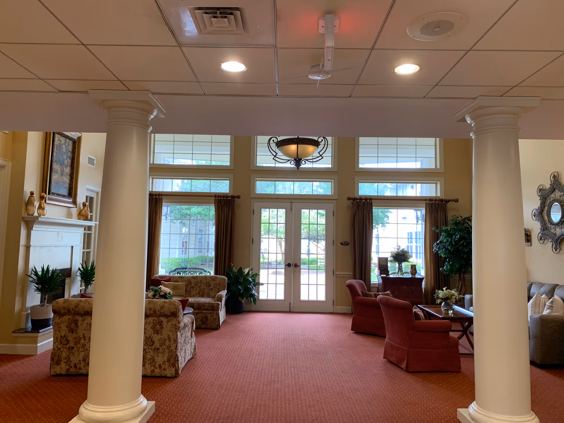 Interior view of a senior living facility common area with large windows and glass doors letting in natural light. The room features two white columns, a red carpet, floral and red upholstered chairs, a fireplace with decorative items, plants, a chandelier, and a decorative mirror on the wall.