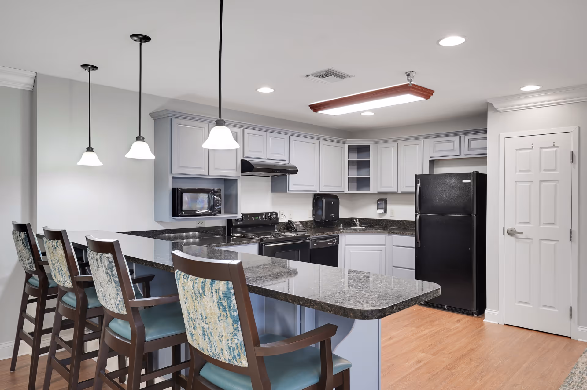 Modern kitchen area with a granite countertop island and four high chairs with patterned backs. The kitchen features white cabinets, a black refrigerator, a microwave, a stove, and a dishwasher. Three pendant lights hang above the island, and the floor is wood.