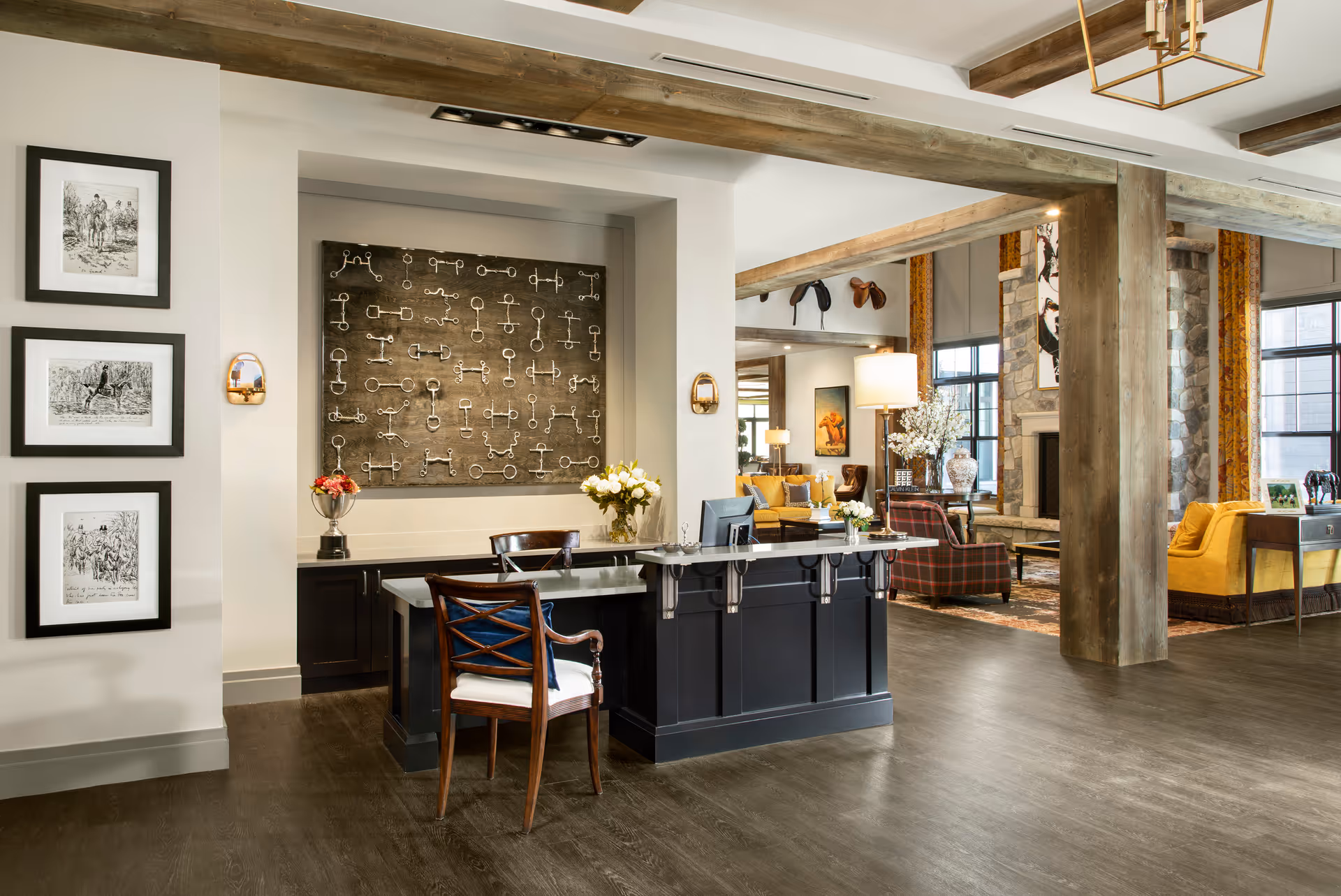 A spacious and well-lit reception area with a dark wood desk and a wooden chair with a white cushion. Behind the desk is a wall decorated with a large artwork featuring various metal keys. To the left, three framed black and white pictures hang on the wall. The room has wooden beams on the ceiling and a polished dark wood floor. In the background, there is a cozy seating area with yellow and plaid armchairs, a stone fireplace, and large windows with yellow curtains.