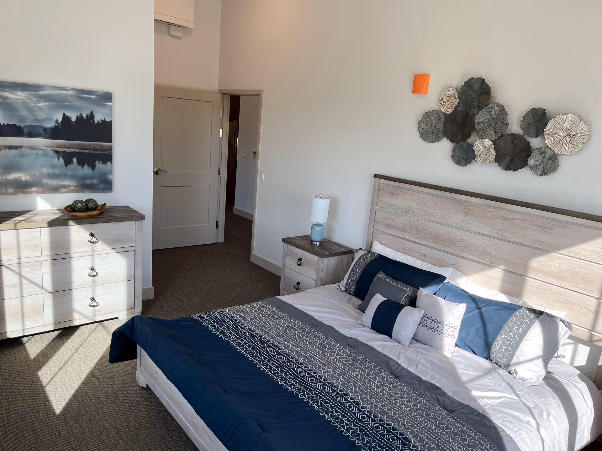 A bright bedroom with a large bed featuring blue and white patterned bedding and multiple pillows. There is a wooden headboard and a matching nightstand with a blue lamp. On the opposite wall, there is a wooden dresser with decorative items and a landscape painting above it. The room has beige carpet and white walls, with sunlight streaming in through a window.