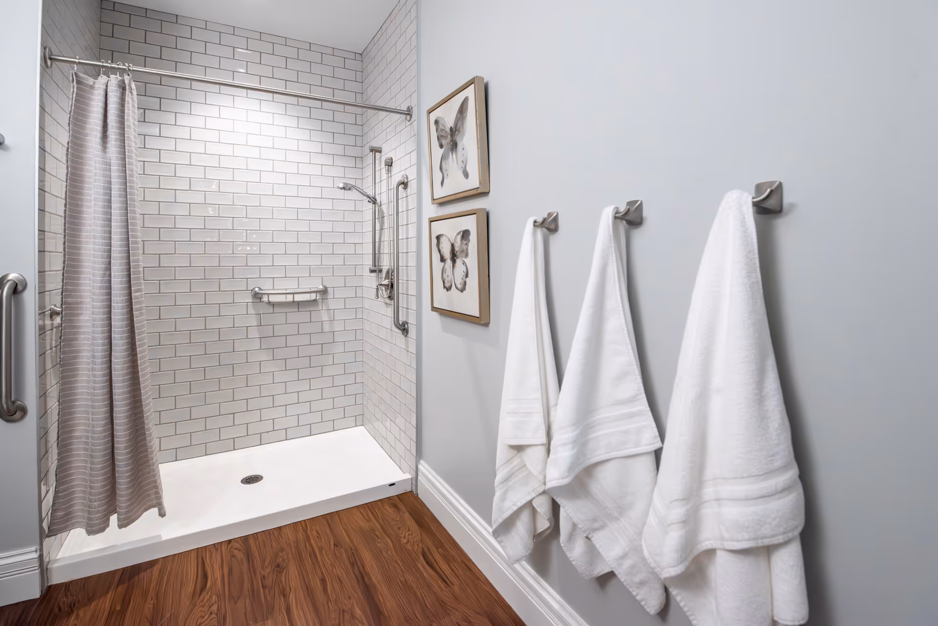 Accessible bathroom with a tiled walk-in shower, grab bars, a striped shower curtain, and three white towels hanging on hooks.