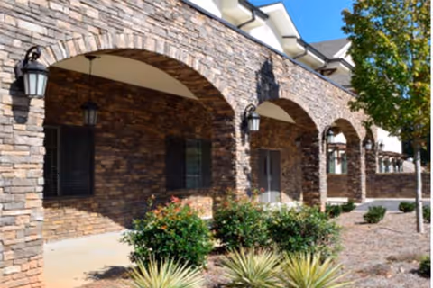 Exterior view of a building with stone archways and lantern-style wall lights. There are shrubs and small plants in front of the building, and a tree on the right side under a clear blue sky.