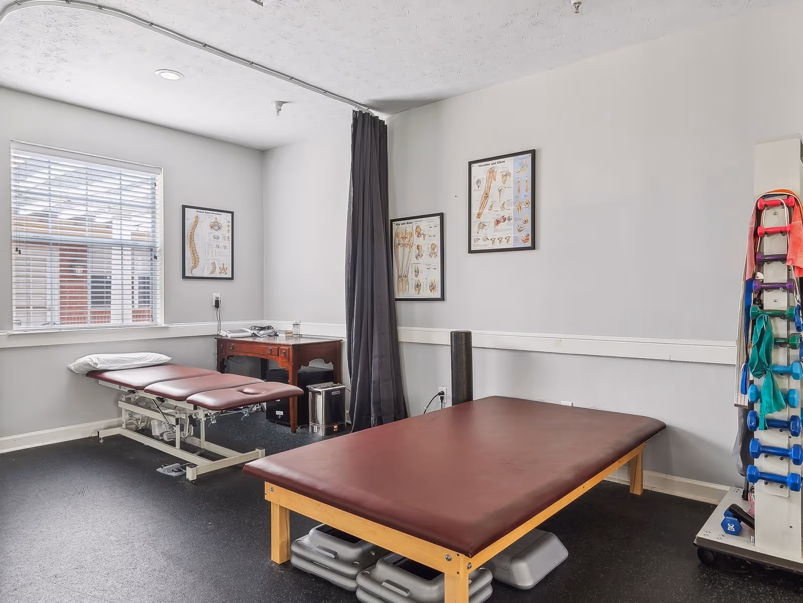 A physical therapy or rehabilitation room with two padded treatment tables, anatomical charts on the walls, a window with blinds, a small wooden desk with medical supplies, and a rack holding colorful resistance bands and small weights.