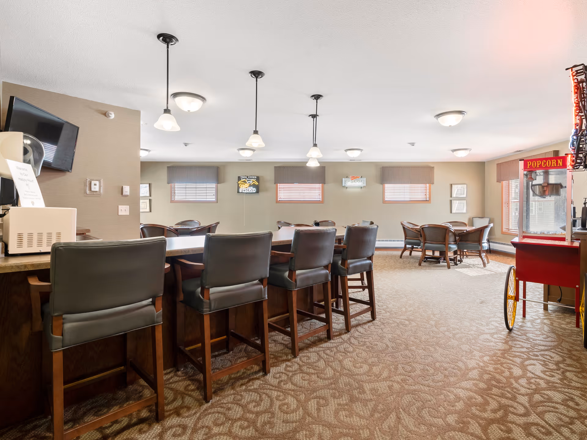 Spacious senior living common dining area with bar seating, tables, pendant lights, and a popcorn cart by the window.