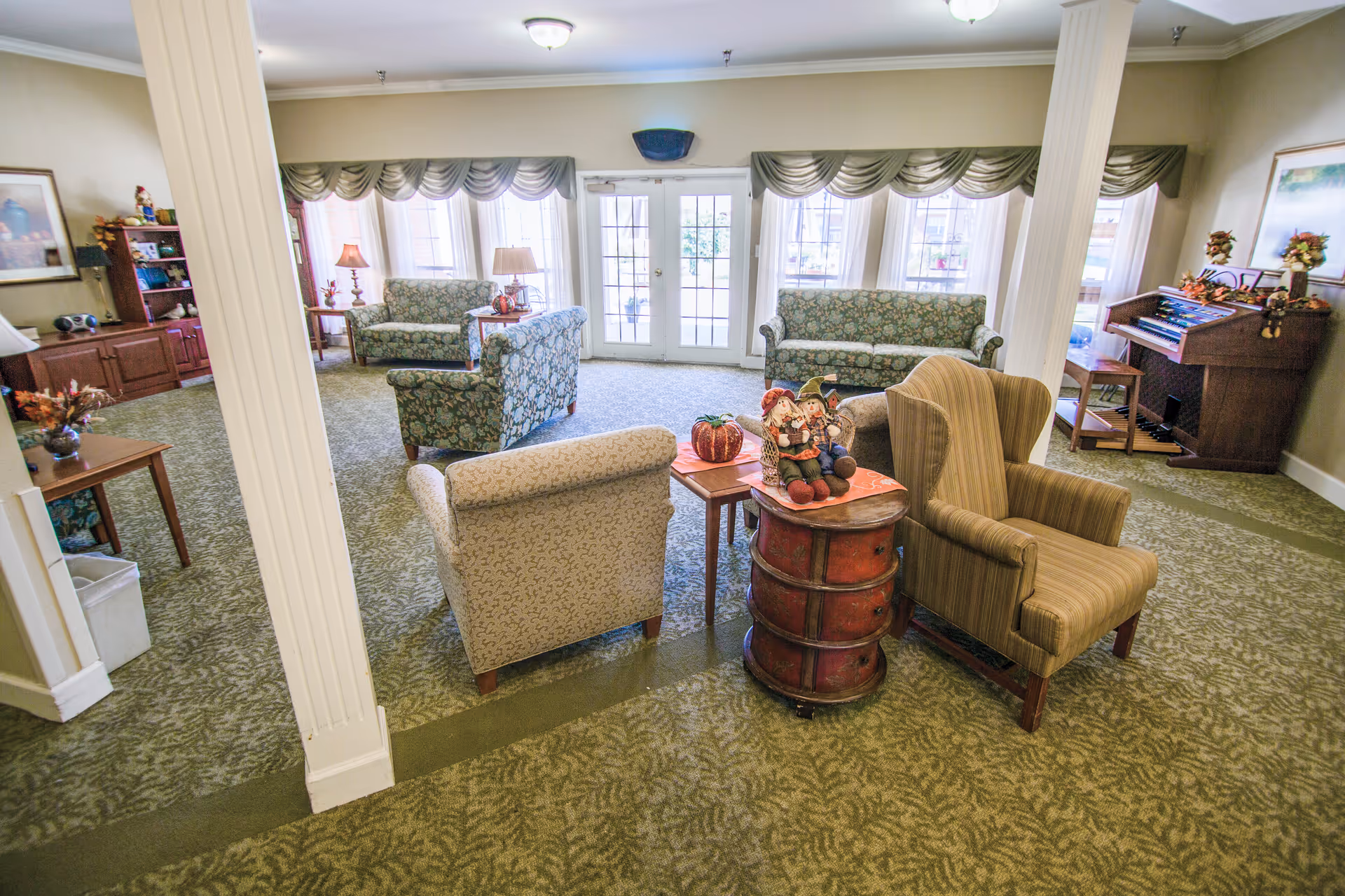 A cozy senior living facility common area with patterned armchairs and sofas arranged around small tables. The room has large windows with green valances, a green patterned carpet, and decorative items including a pumpkin and scarecrow dolls on a small round table. There is an organ in the corner and framed artwork on the walls.