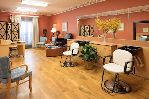 Interior of a salon area in a senior living facility with wooden flooring, two white salon chairs in front of a long mirror, a wooden counter with a vase of yellow flowers, a potted plant on the floor, and additional seating including blue and wooden chairs. The walls are painted red with a decorative border near the ceiling and a window with curtains in the background.
